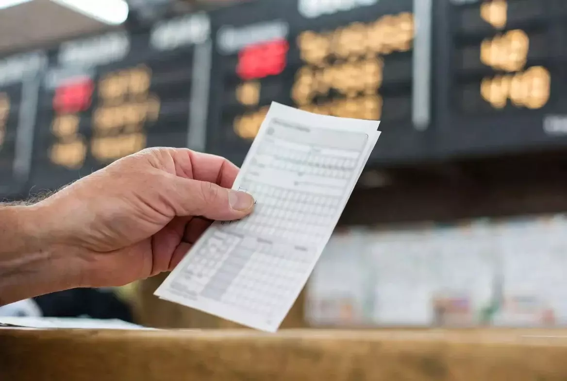 Hand hält einen Wettschein vor einer Anzeigetafel mit Quoten